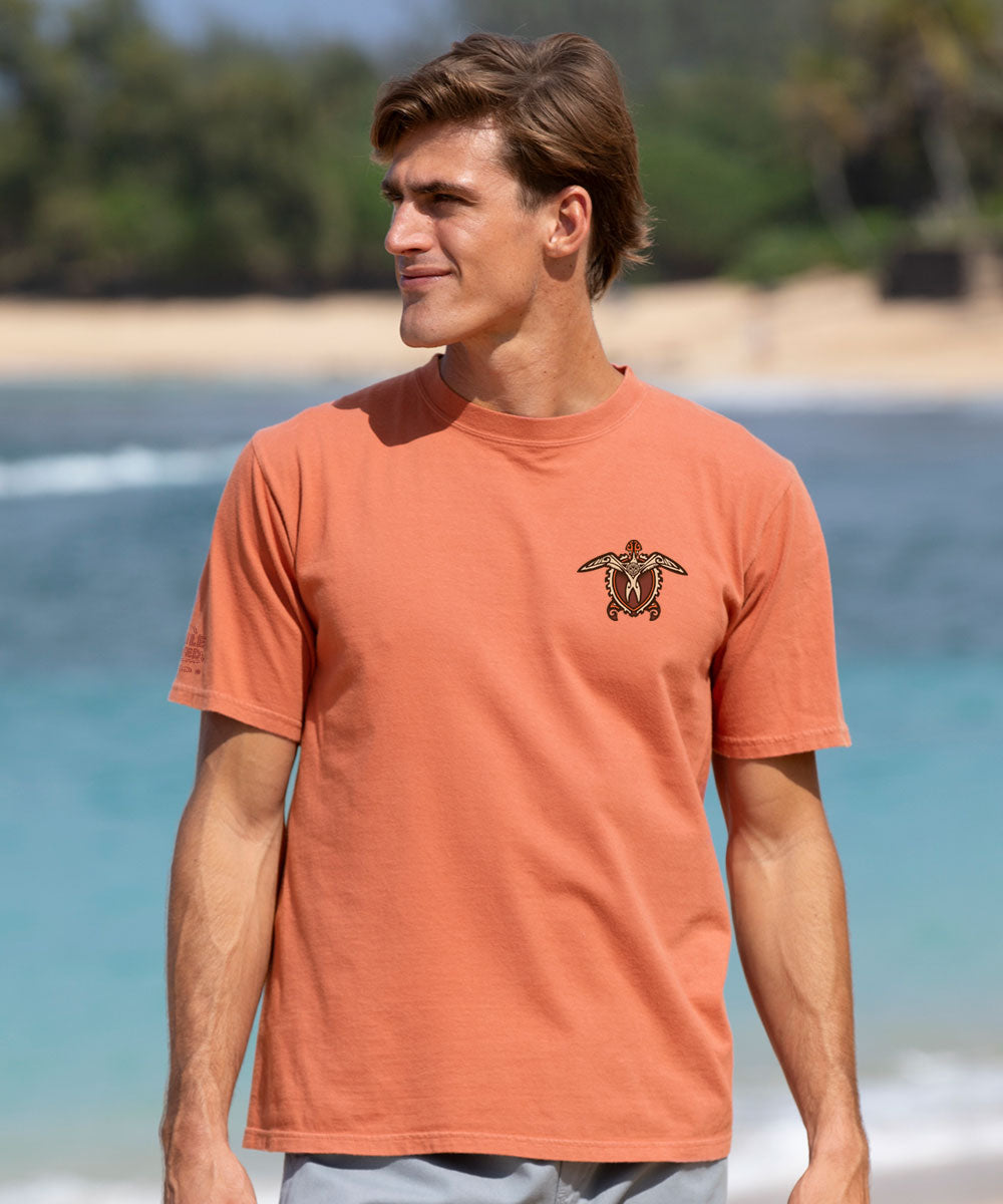 A man wearing a orange color t-shirt with a small tribal turtle graphic on the left chest, standing in front of the ocean.  View 3