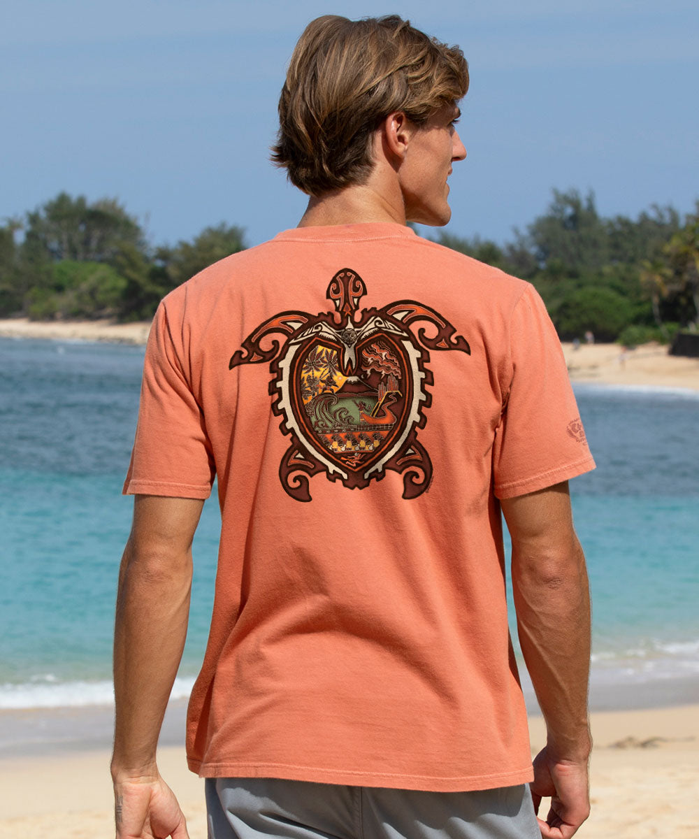 A man standing on a beach, viewed from behind, wearing a coral orange t-shirt with a large detailed turtle graphic printed on the back. View 2