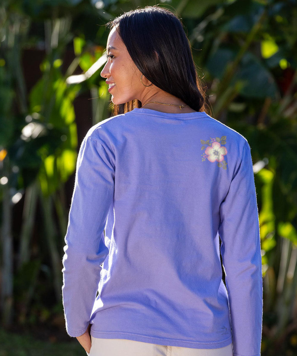 A woman shown from the back wearing a lavender long-sleeve shirt with a small white and yellow floral graphic on the upper right back, set against a background of green tropical foliage. View 3
