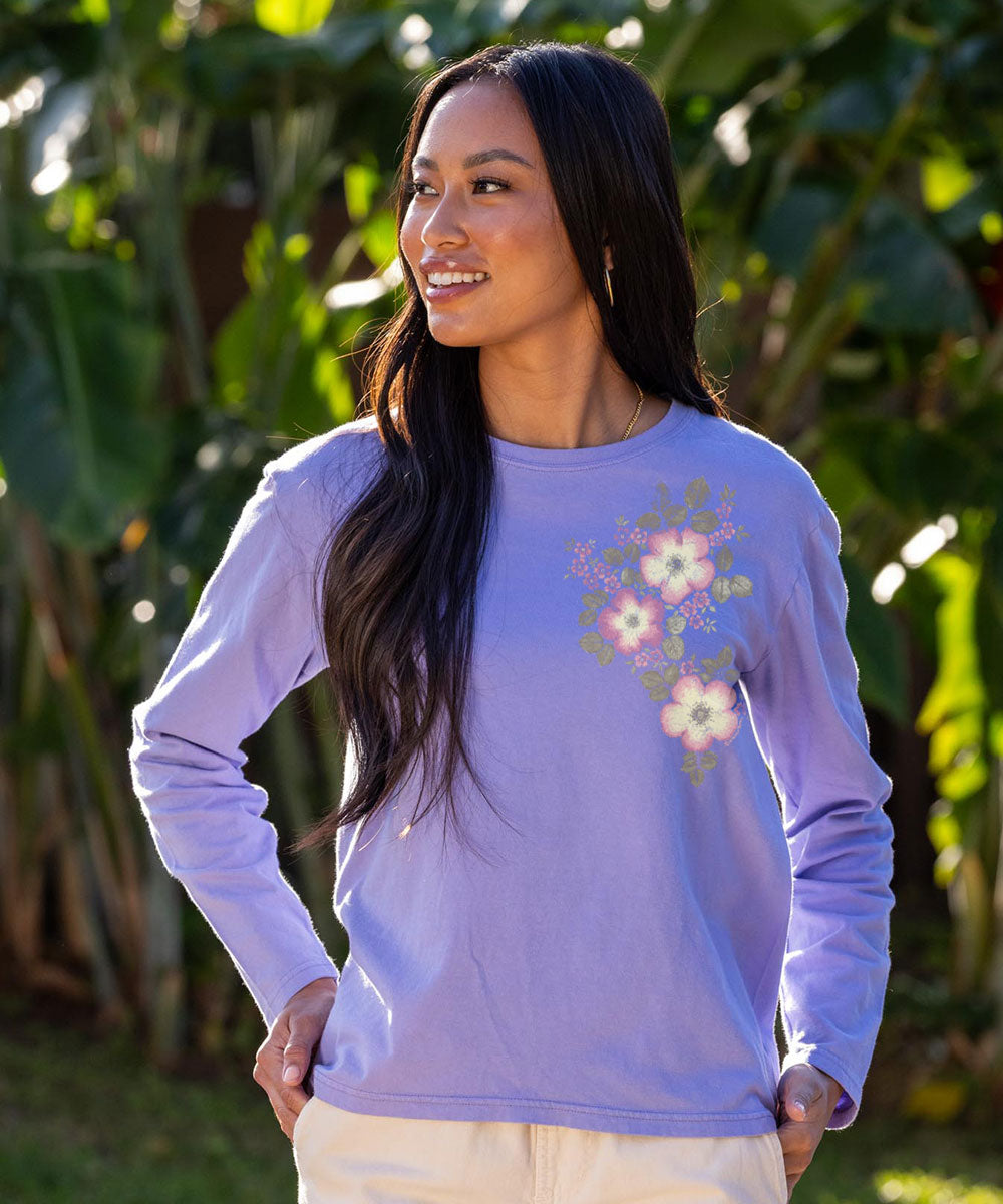 A woman wearing a lavender long-sleeve crewneck shirt with a floral graphic on the left chest, standing outdoors in front of lush green tropical foliage. View 2