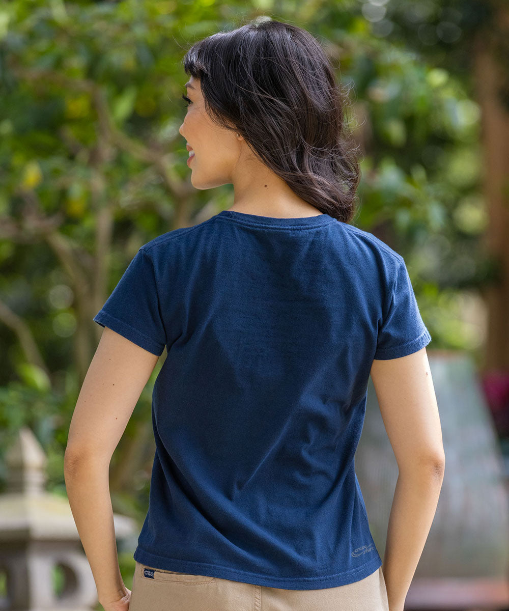 A woman wearing a dark navy blue short-sleeve t-shirt, shown from the back in an outdoor setting with green foliage in the background. View 3