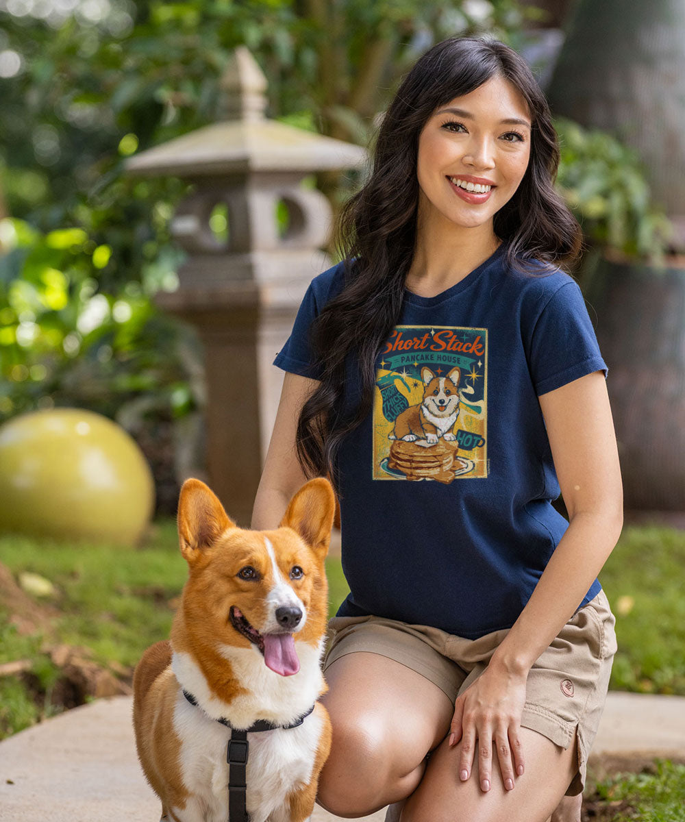 A woman kneeling outdoors next to a corgi dog, wearing a navy blue graphic t-shirt with a vintage-style 'Just Shit' cartoon dog print and khaki shorts. View 2