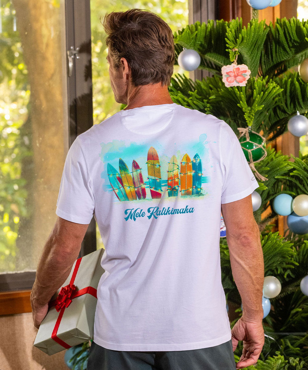 A man viewed from behind wearing a white t-shirt with a colorful graphic of surfboards and the text 'Wai Kalihomuka' on the back. He is holding a wrapped gift box next to a decorated Christmas tree. View 2