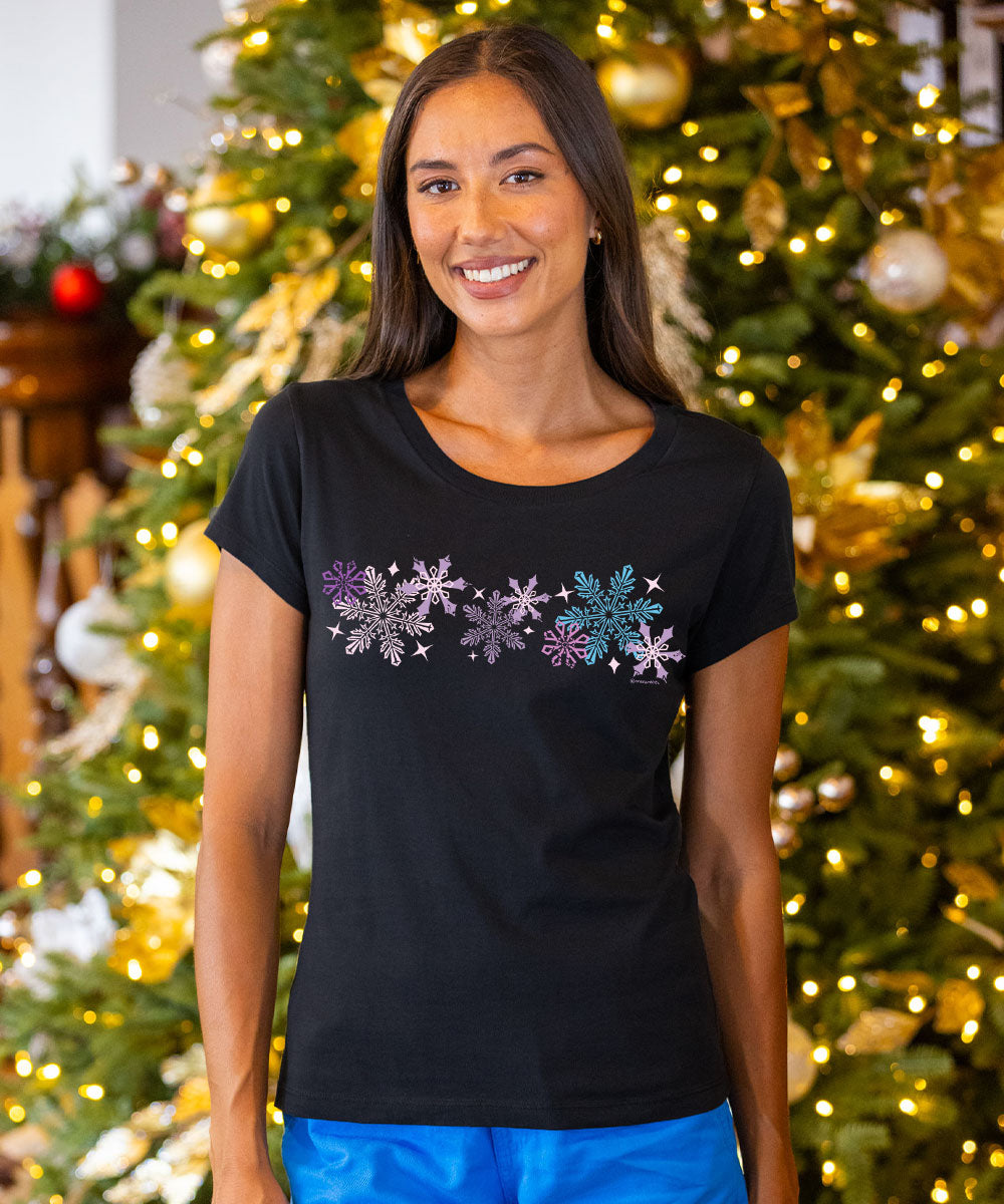 A woman wearing a black short-sleeve t-shirt with a row of colorful glittery snowflakes across the chest, standing in front of a decorated Christmas tree. View 2