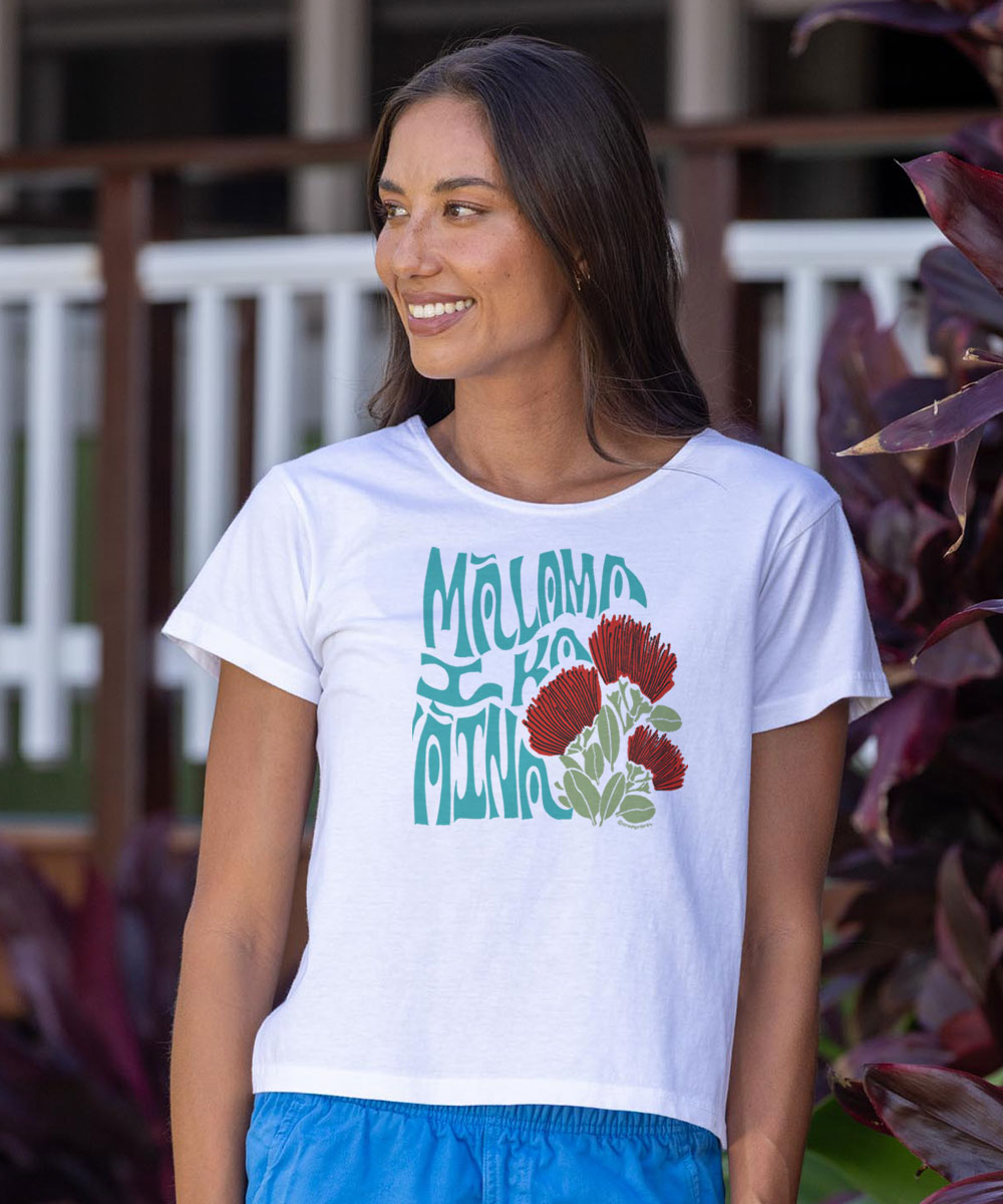 A woman wearing a white short-sleeve graphic t-shirt with teal lettering alongside red and green flowers, set against a tropical outdoor background. View 2