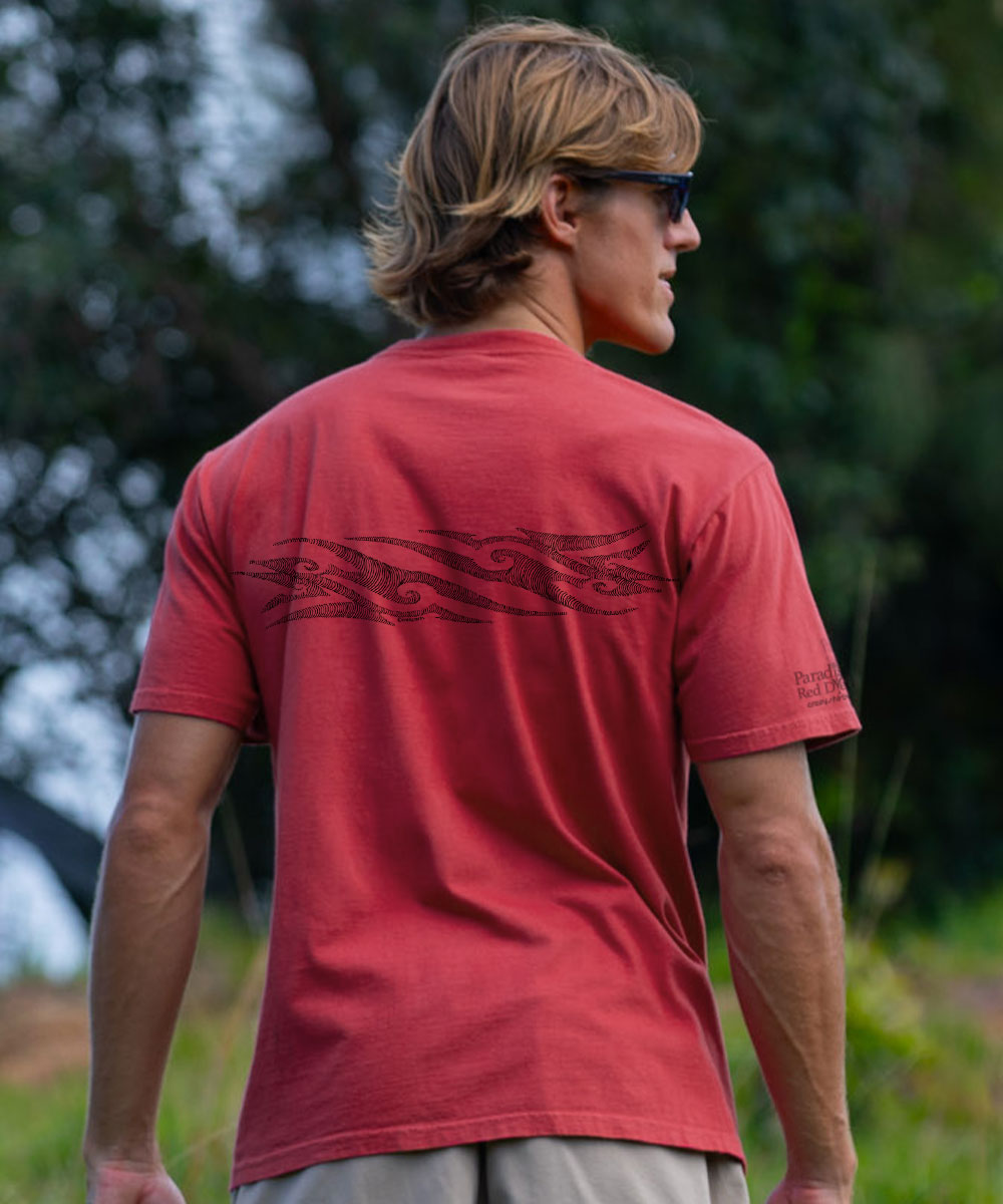 A man wearing a red short-sleeve t-shirt with a black tribal-style graphic across the upper back, photographed outdoors with a blurred green background. View 3