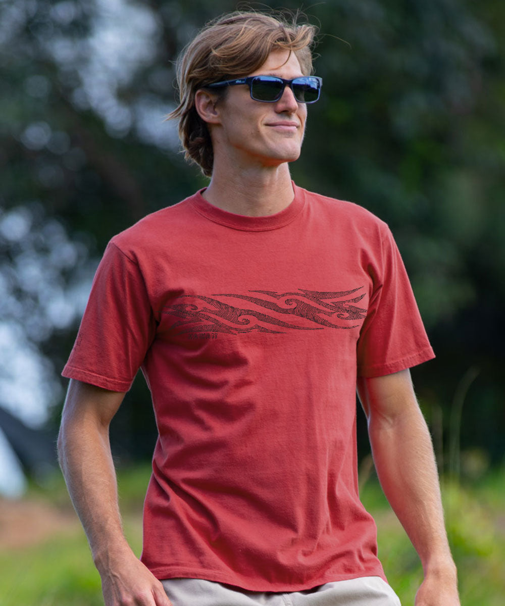 A man wearing a red crewneck t-shirt with a dark tribal-style graphic across the chest, set against a blurred outdoor background. View 2