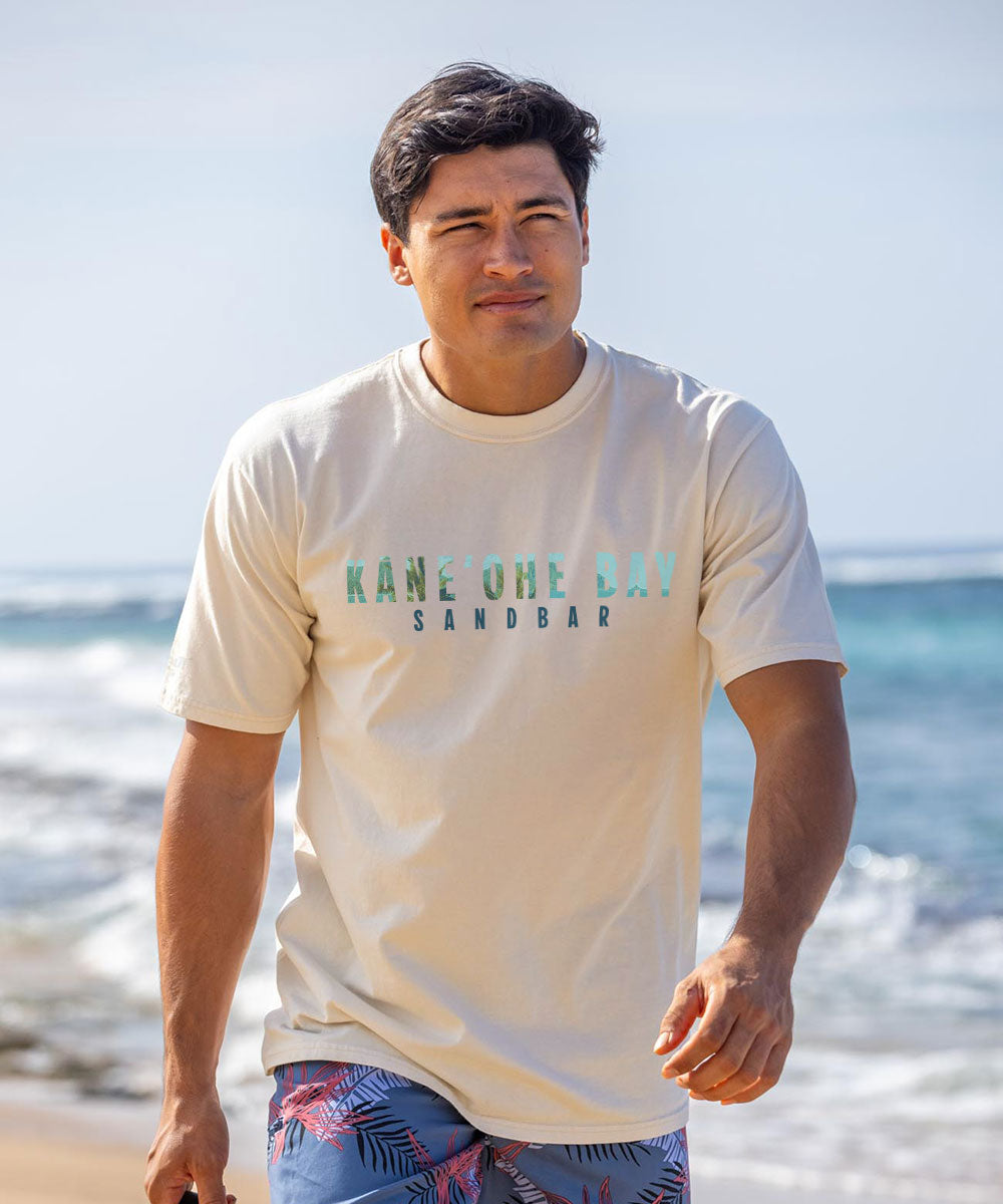 A man wearing a cream t-shirt that says "Kane'ohe Bay" walking on the beach.  View 3