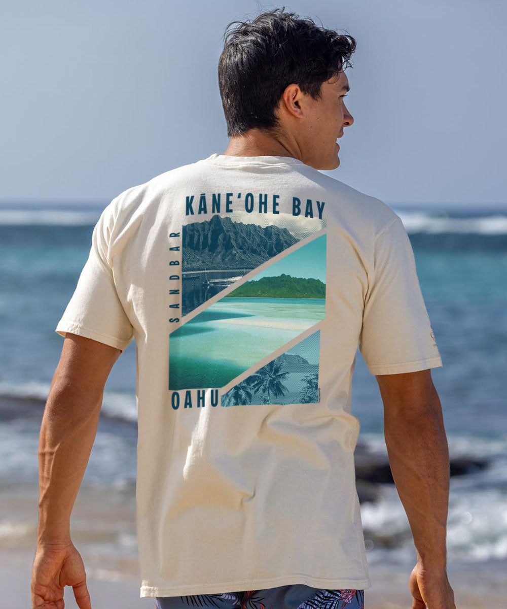A man wearing a cream-colored t-shirt with a large graphic print of Kane'ohe Bay, Oahu, shown from the back in an outdoor beach setting. View 2