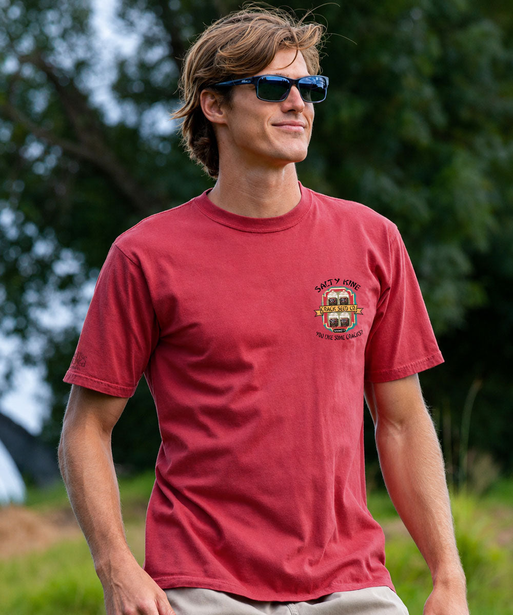 A man wearing a red crewneck t-shirt with a small chest graphic and sunglasses, standing outdoors against a blurred green background. View 3
