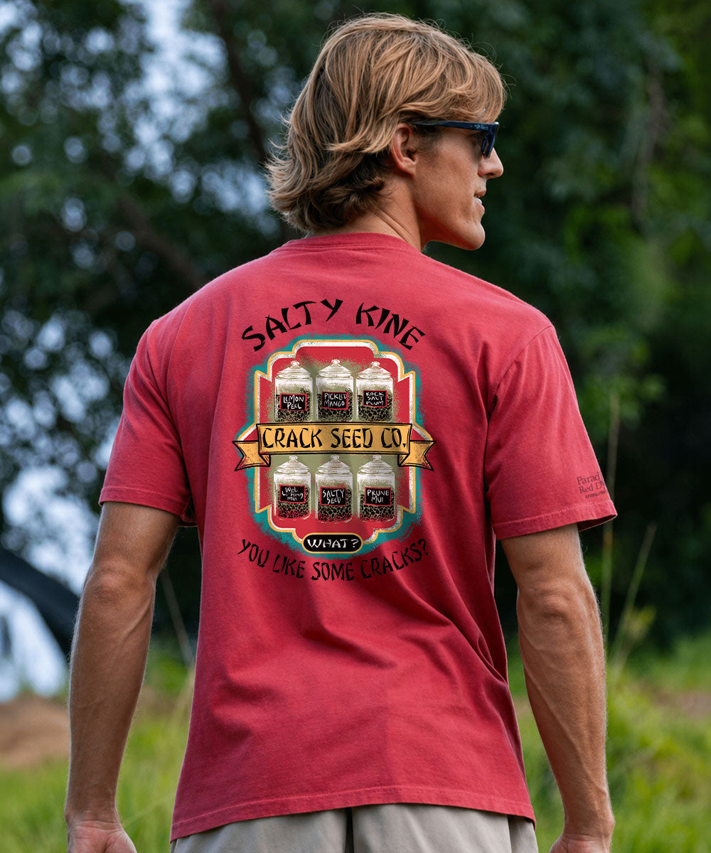 A man wearing a red graphic t-shirt with a large back print reading 'SALTY KINE' and 'CRACK SEED CO.' is shown from behind in an outdoor setting with trees in the background. View 2