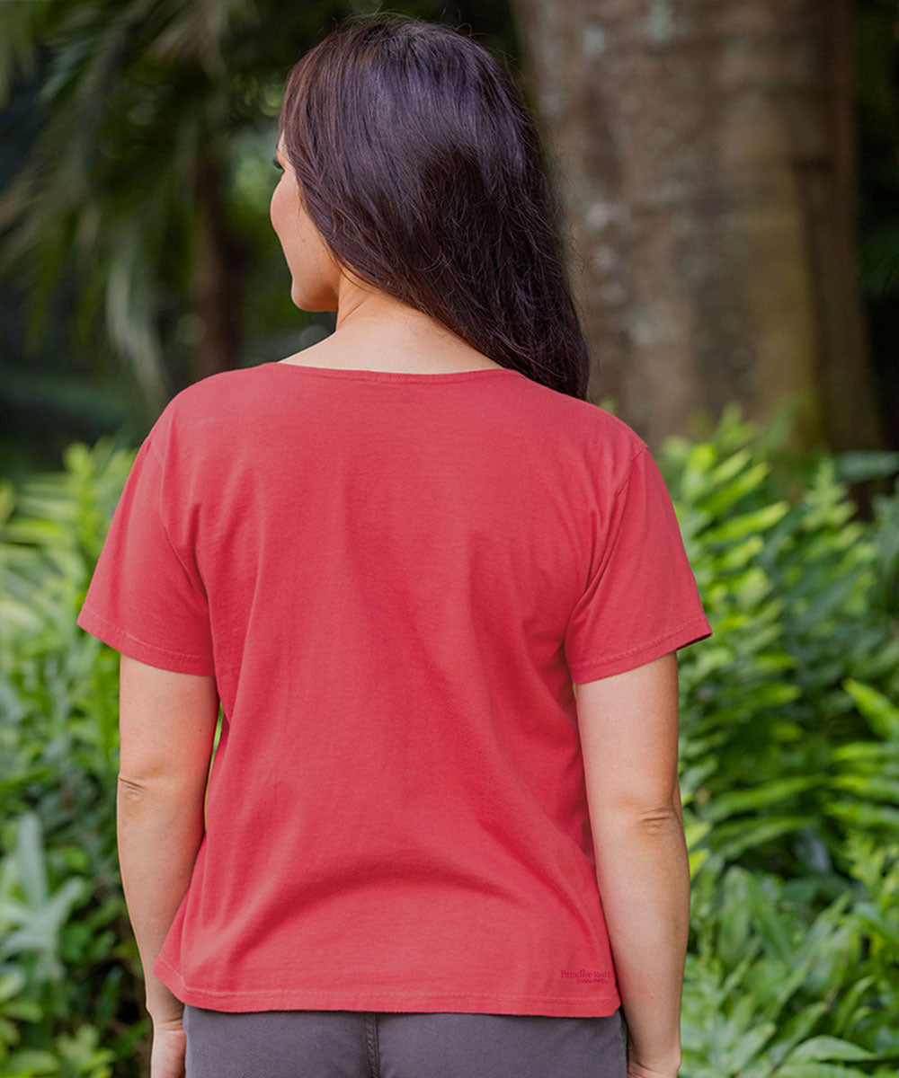 A woman viewed from behind wearing a solid red short-sleeve t-shirt and dark gray pants, standing outdoors in front of lush green foliage. View 3