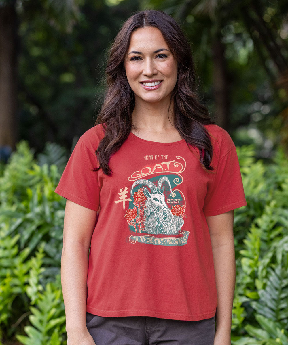 A woman wearing a red graphic t-shirt with a goat design and text, standing outdoors in front of a blurred green foliage background. View 2