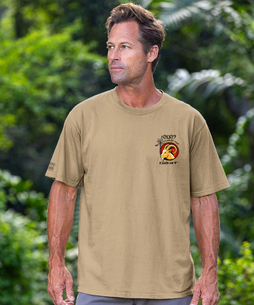 A man wearing a tan crewneck t-shirt with a small graphic on the left chest, standing outdoors in front of a blurred green foliage background. View 3