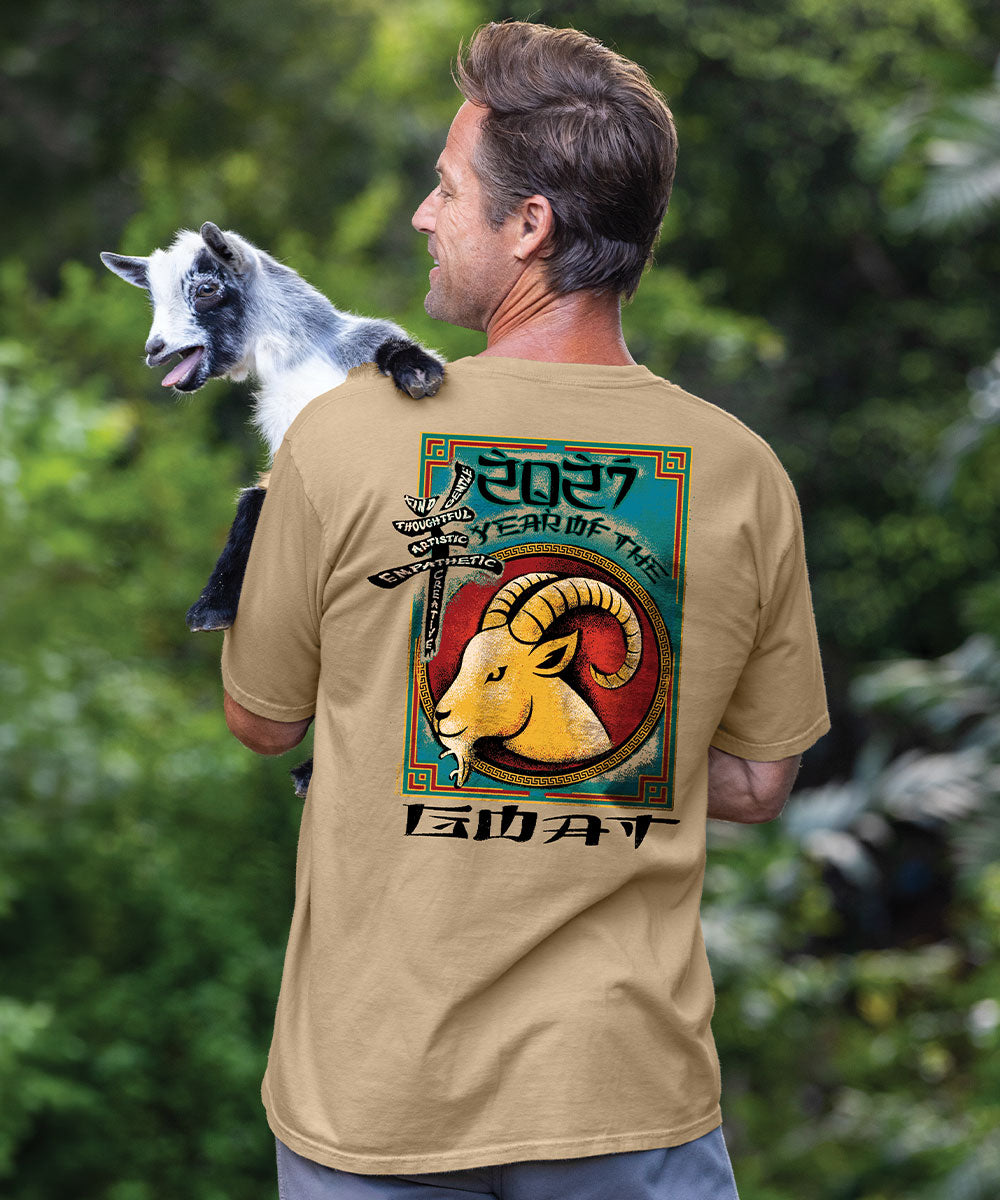 A man wearing a tan graphic t-shirt with a '2027 Year of the Goat' design on the back, holding a small goat in his arms against a green forest background. View 2