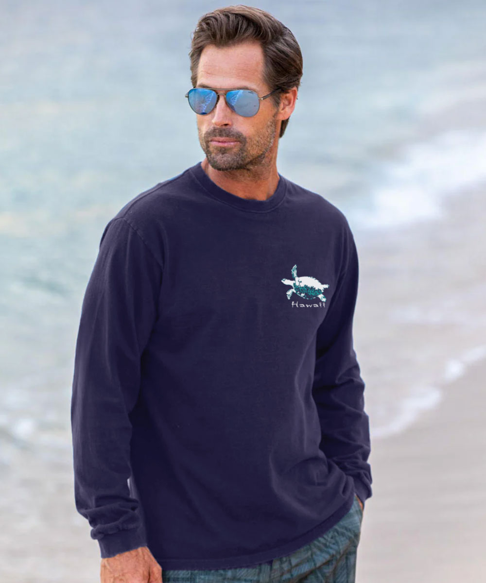 A man wearing a navy blue long-sleeve crewneck shirt with a small turtle graphic on the left chest, standing on a beach with the ocean in the background. View 3