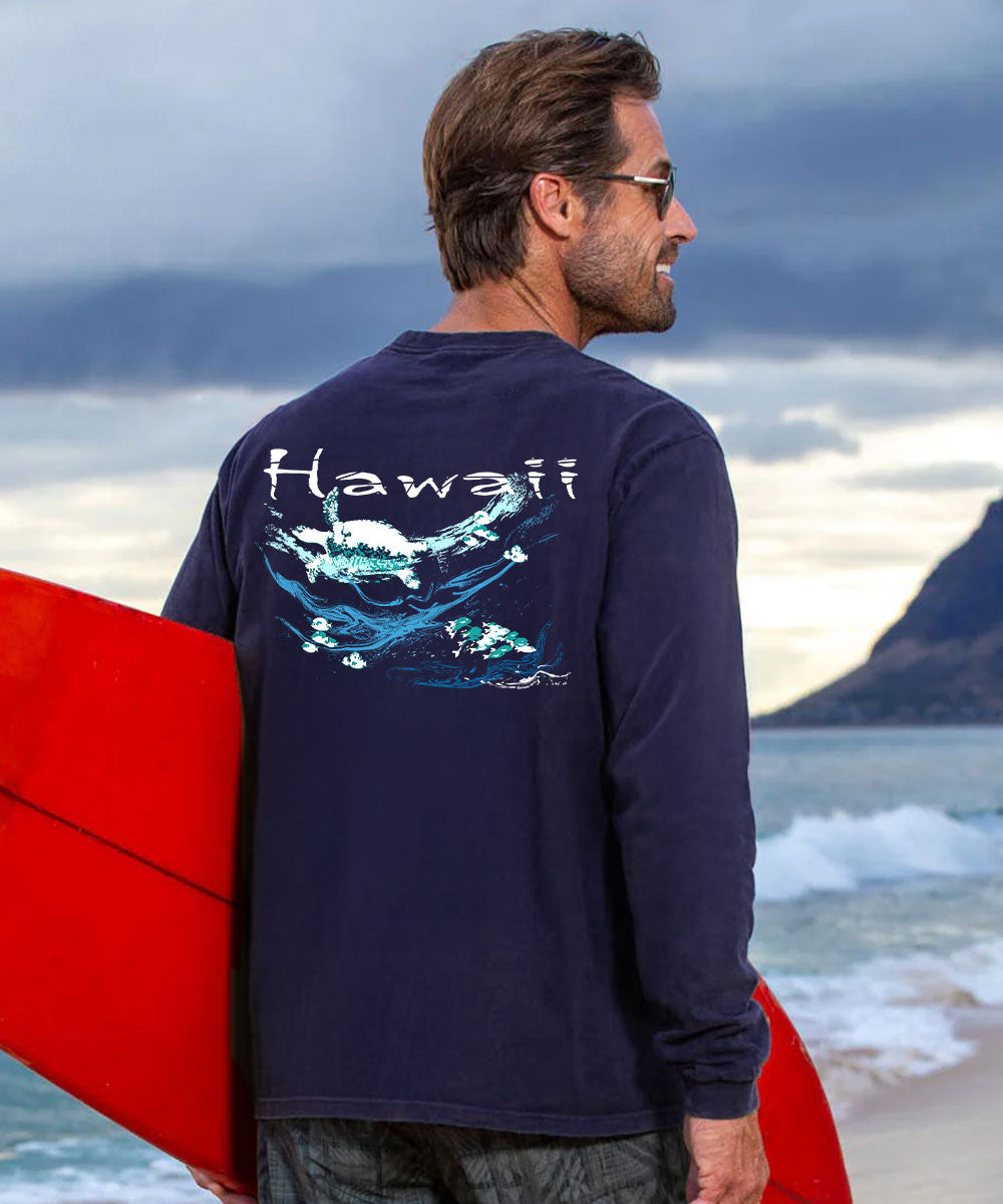 A man on a beach wearing a navy blue long-sleeve shirt with a large graphic print on the back that reads 'Hawaii' above blue waves and fish, holding a red surfboard. View 2