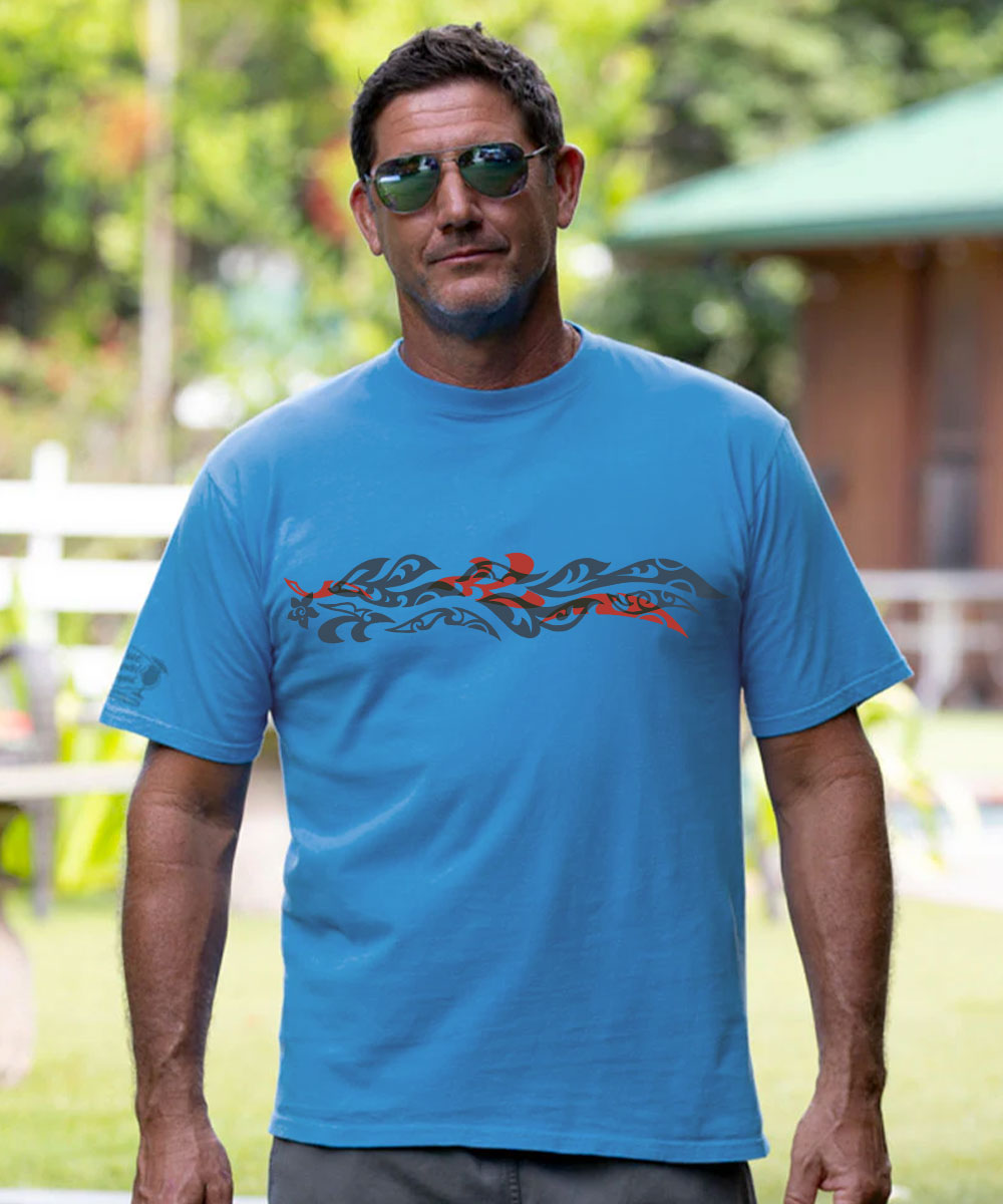 A man wearing a bright blue short-sleeve t-shirt with a black and red Polynesian tribal graphic across the chest, standing outdoors in a sunny, green environment. View 2