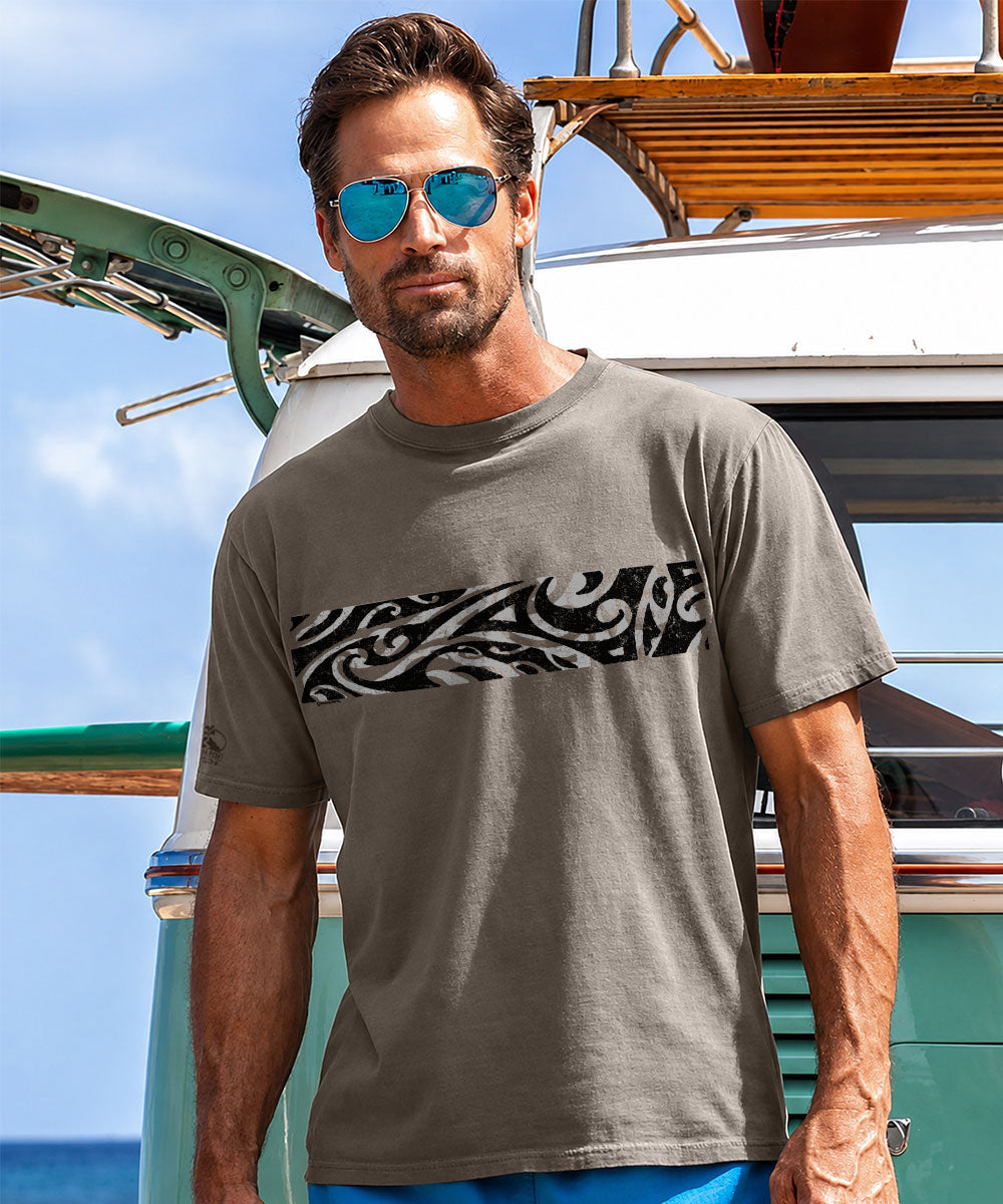 A man wearing a taupe gray crewneck t-shirt with a black and white Polynesian wave pattern graphic across the chest, standing in front of a van. View 2