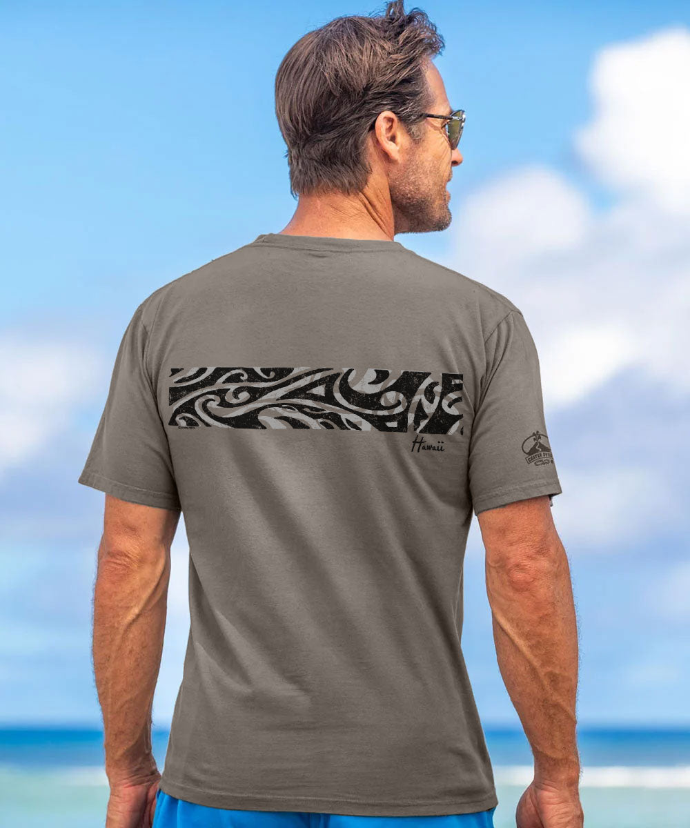 A man wearing a gray short-sleeve t-shirt with a black and white Polynesian-style wave graphic across the upper back, photographed from behind against a beach background. View 3