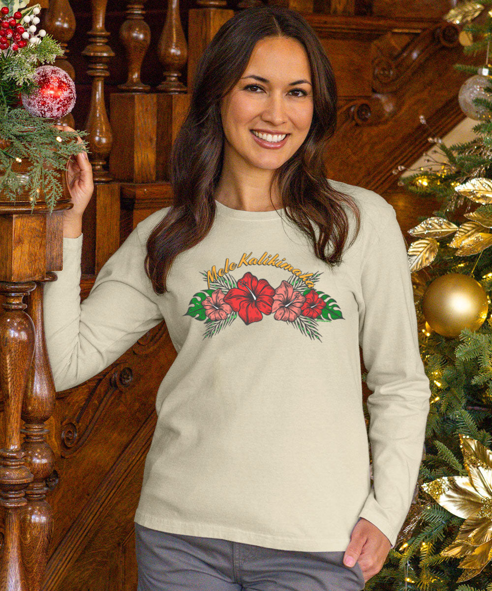 A woman wearing a light beige long-sleeve t-shirt with a colorful 'Hula Dancer' graphic featuring hibiscus flowers, posed in front of a wooden staircase and a decorated Christmas tree.