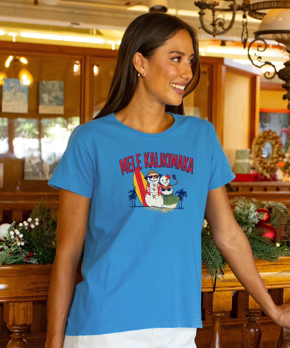A woman wearing a bright blue graphic t-shirt with the Hawaiian text and a cartoon snowman design, standing in a warm, wood-paneled indoor setting. View 2