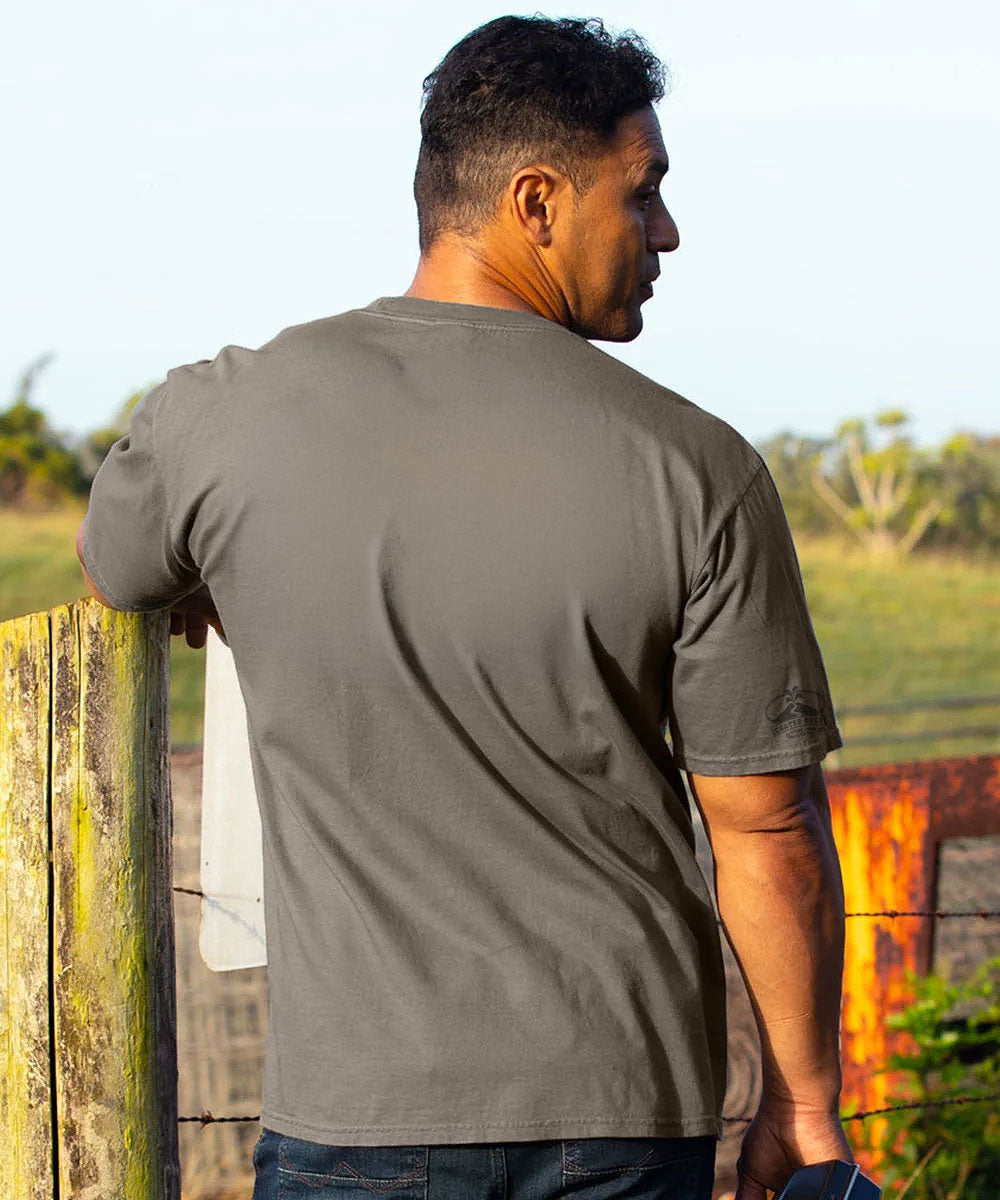 A man wearing a dark gray crewneck t-shirt, viewed from behind as he leans against a weathered wooden fence in an outdoor rural setting. View 3