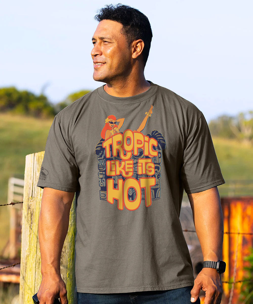 A man wearing a dark gray graphic t-shirt with a tropical beach-themed print that reads 'TROPIC LIKE TO HOT'. The background is a blurred outdoor scene with a wooden fence and green hills under a clear sky. View 2