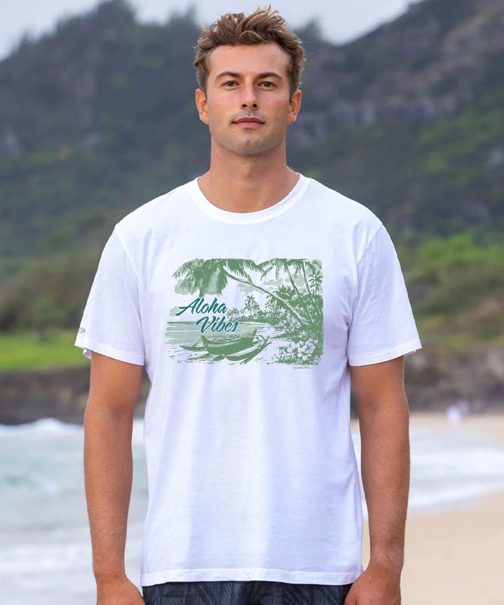 A man standing on a beach wearing a white crewneck t-shirt with a green tropical graphic that reads 'Aloha West'. View 2