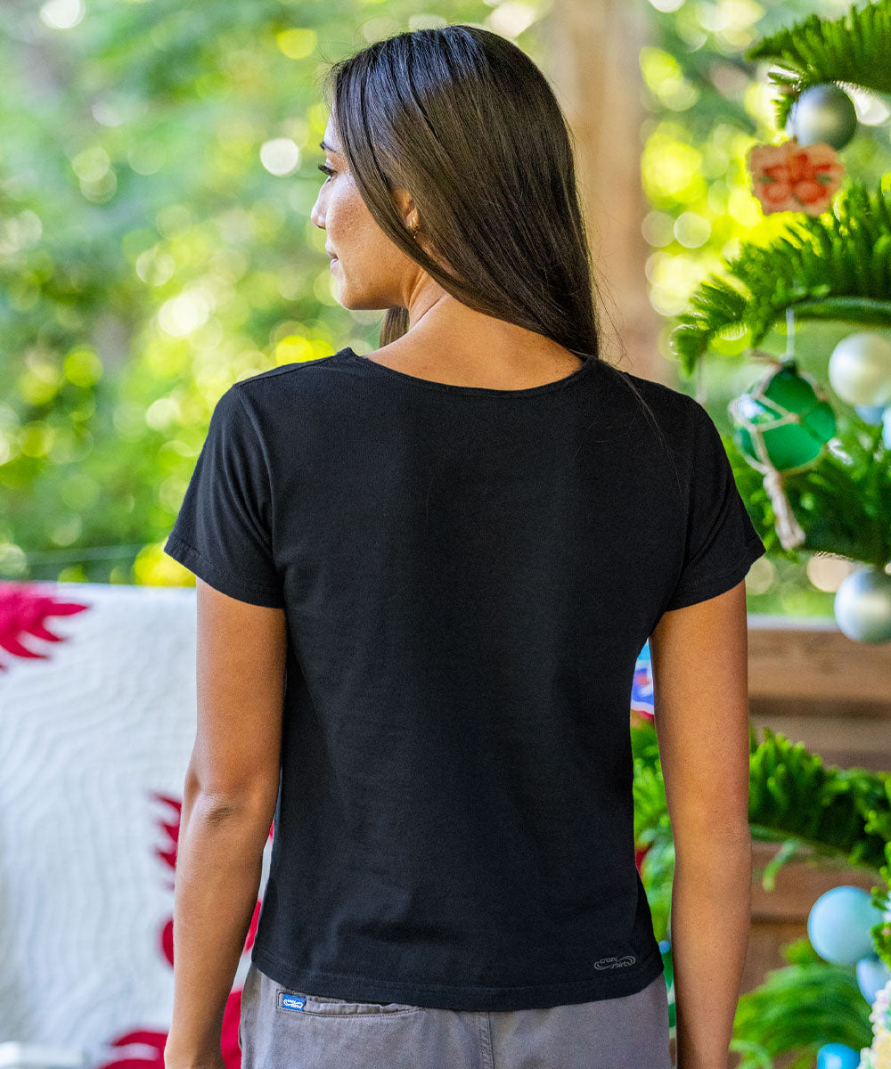 A woman viewed from behind wearing a black short-sleeve crewneck t-shirt, standing in front of a blurred outdoor background with a decorated Christmas tree. View 3