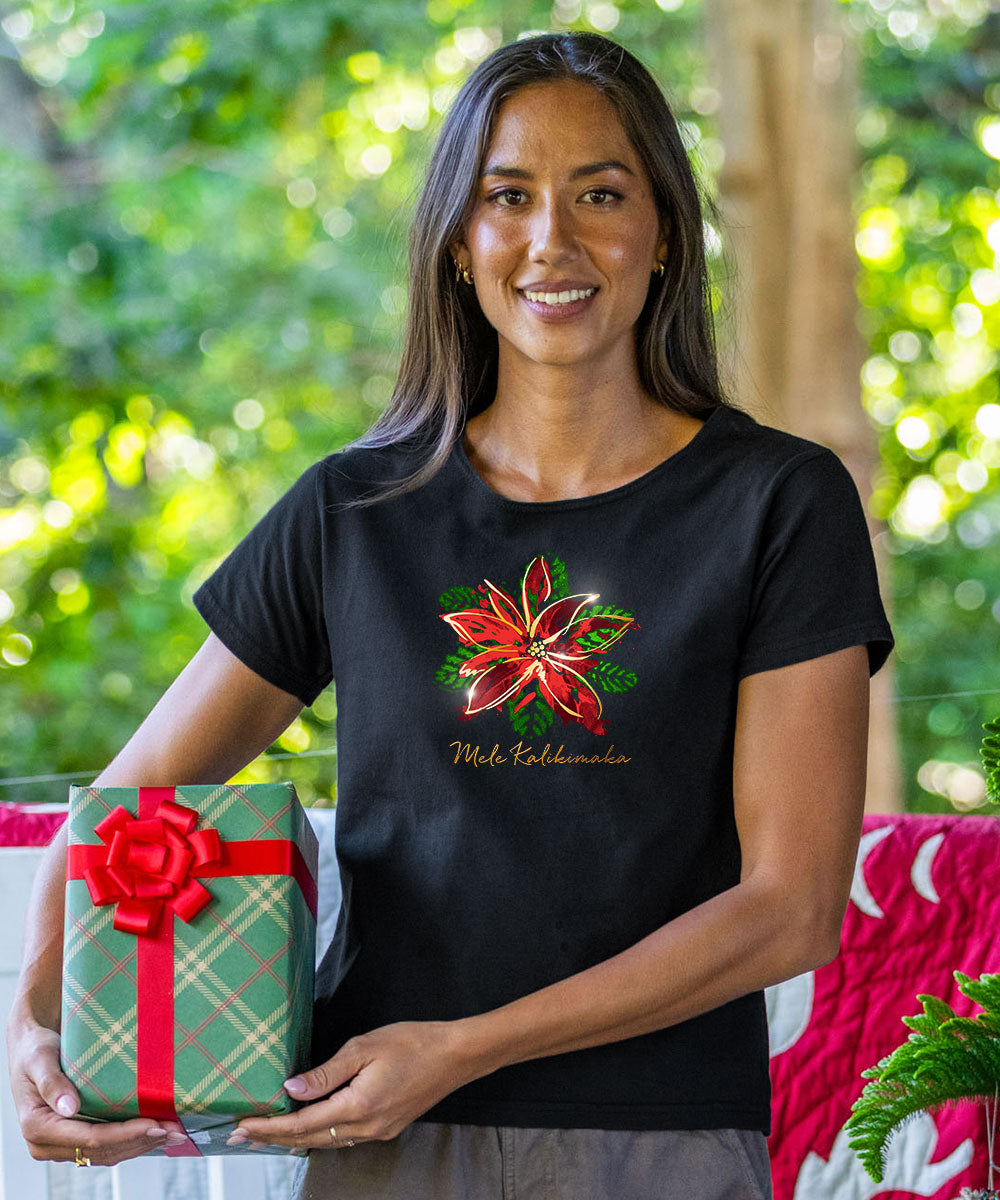 A woman wearing a black t-shirt with a red poinsettia and gold 'Merry Christmas' text, holding a green and red gift box in front of a blurred outdoor garden background. View 2