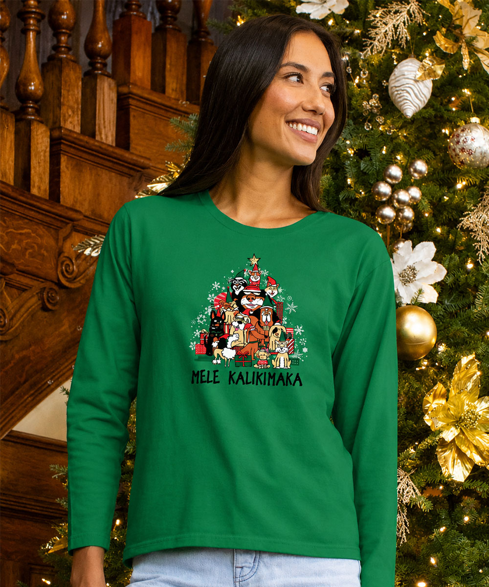 A woman wearing a bright green long-sleeve shirt with a festive Christmas graphic and the text 'MELE KALIKIMAKA' printed on the front, posed in front of a decorated Christmas tree and wooden staircase. View 2