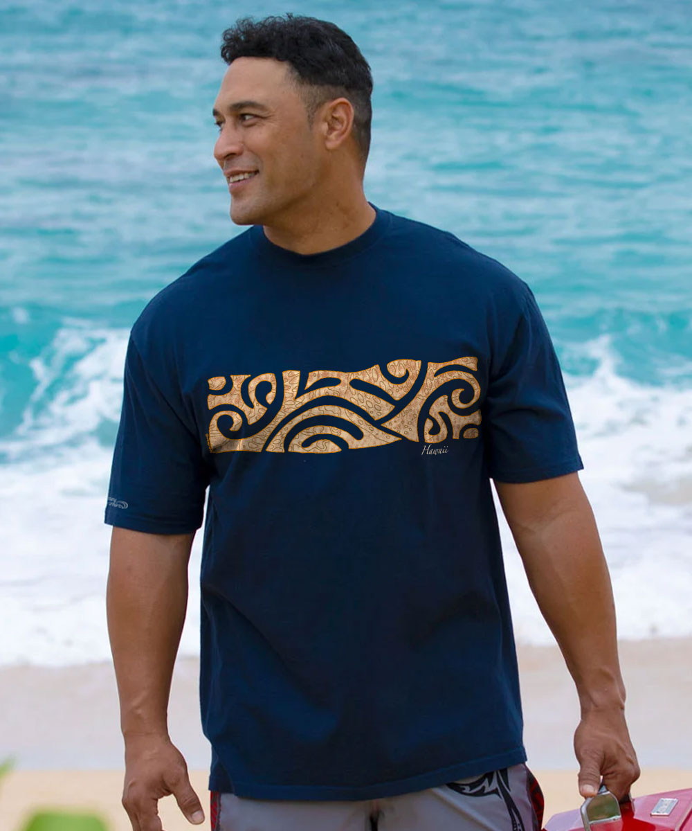 A man wearing a navy blue Hawaii t-shirt with a gold Polynesian tribal pattern across the chest, standing on a beach with the ocean waves in the background. View 2