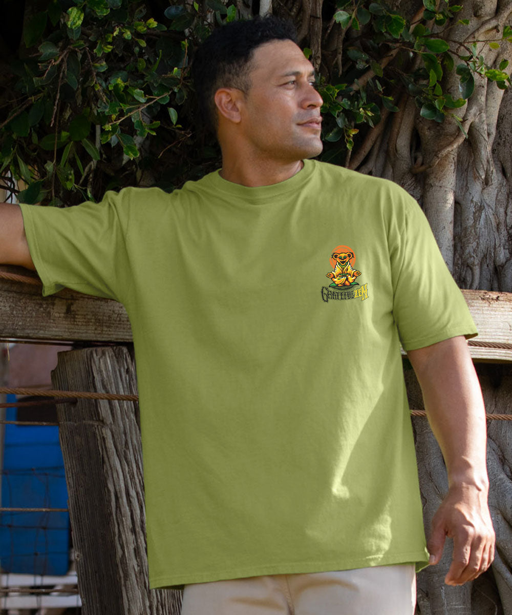 A man wearing a light olive green crewneck t-shirt with a small yellow and black graphic on the left chest, posed outdoors against a rustic wooden fence and tree trunk. View 3