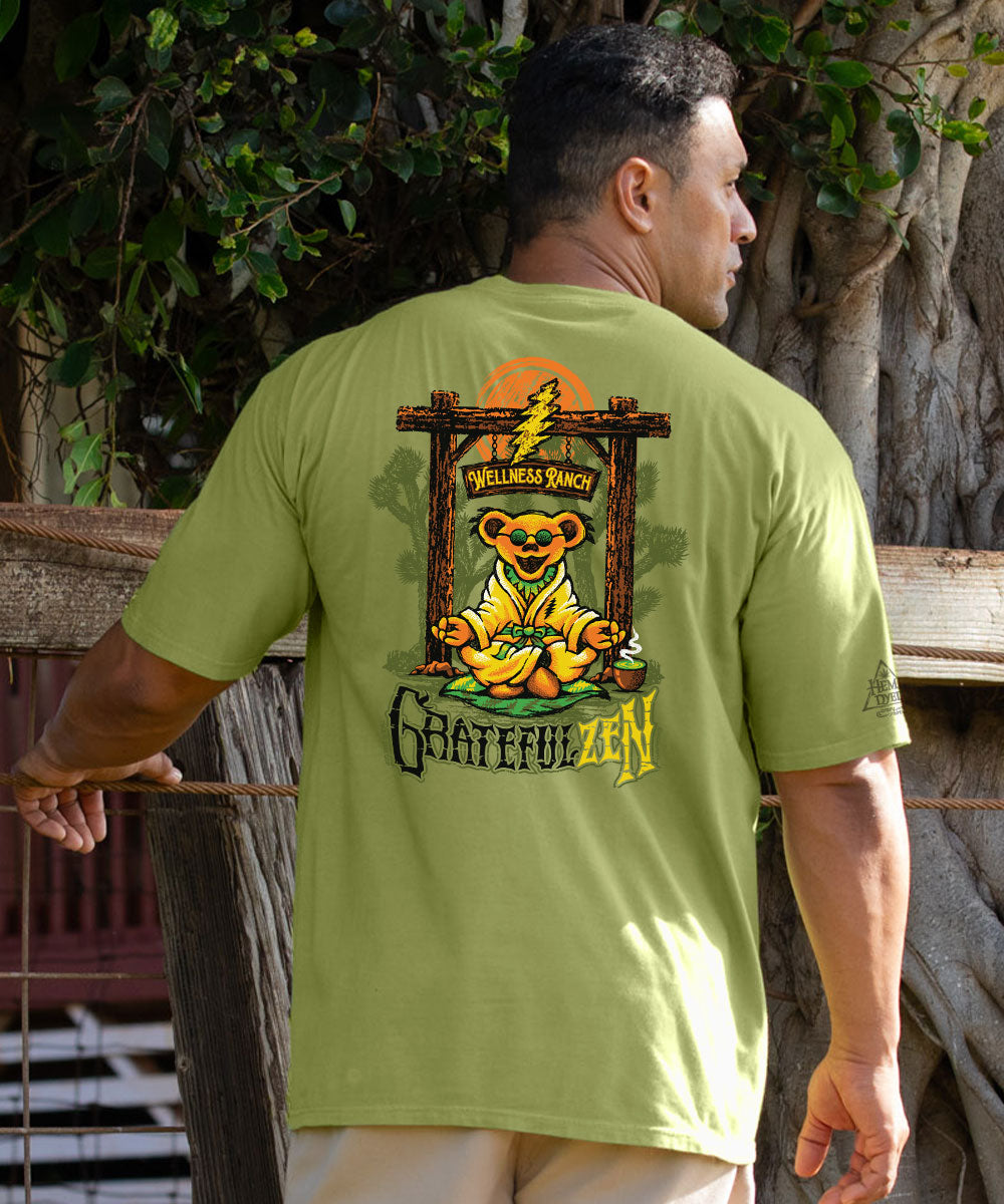 A man wearing a light olive green t-shirt, viewed from behind, showing a large graphic print of a meditating Grateful Dead bear with the words 'Wellness Ranch' and 'Grateful Zen'. View 2