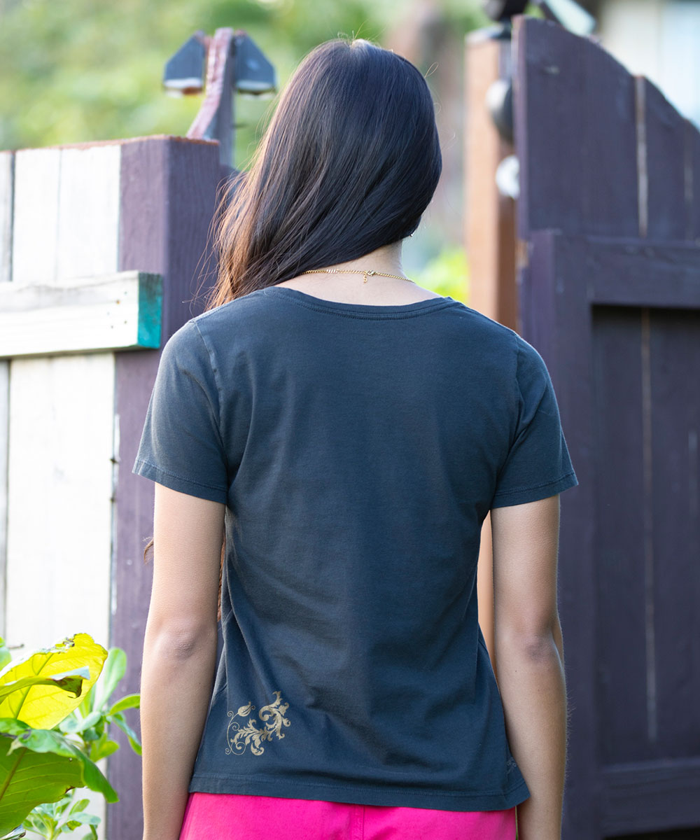 A woman viewed from behind wearing a black t-shirt with a small gold bicycle graphic on the bottom left, paired with bright pink pants, standing outdoors in front of a wooden fence. View 3