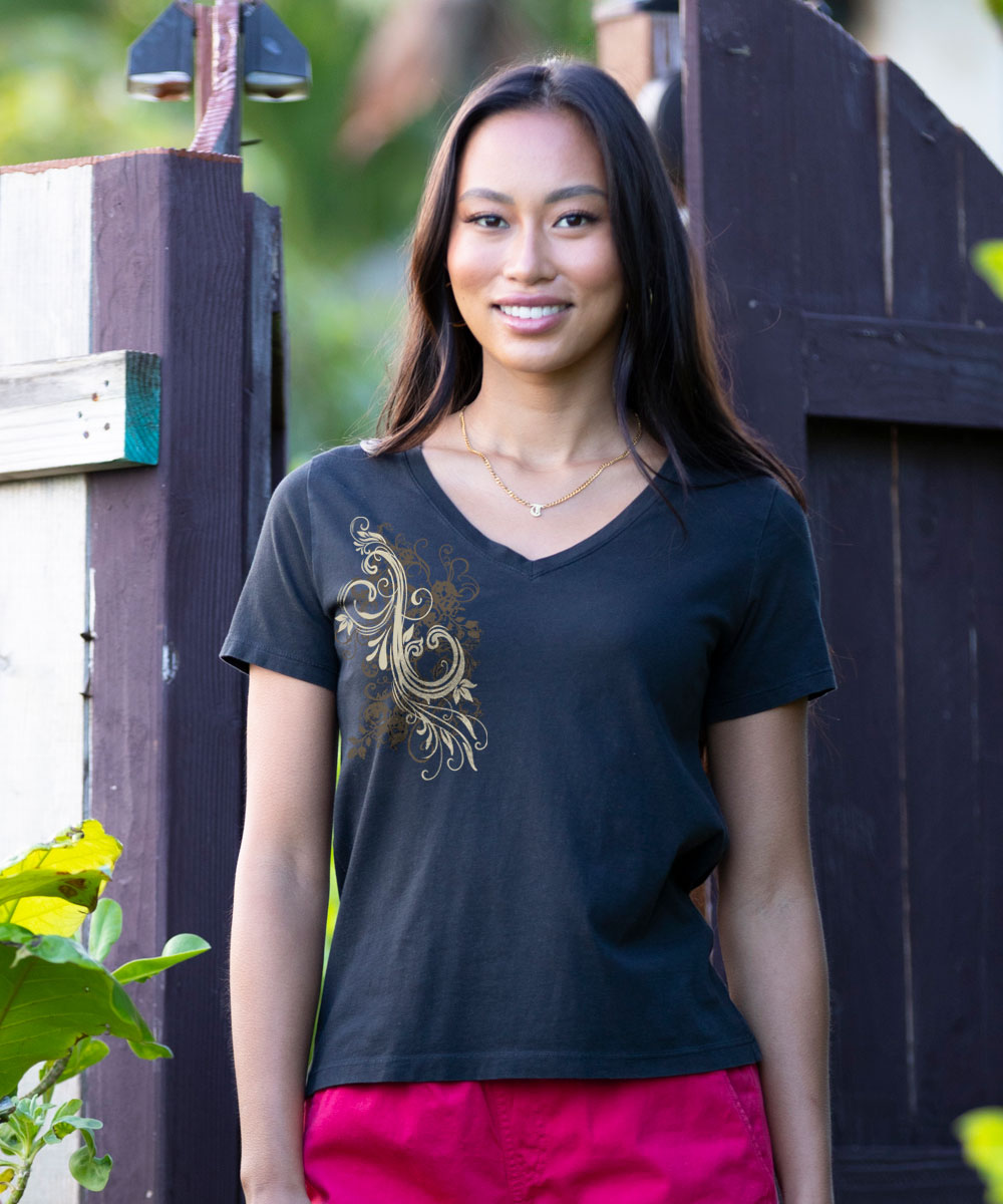 A woman wearing a black V-neck t-shirt with a gold baroque scroll graphic on the left chest, paired with bright pink pants, standing in front of a dark wooden gate. View 2