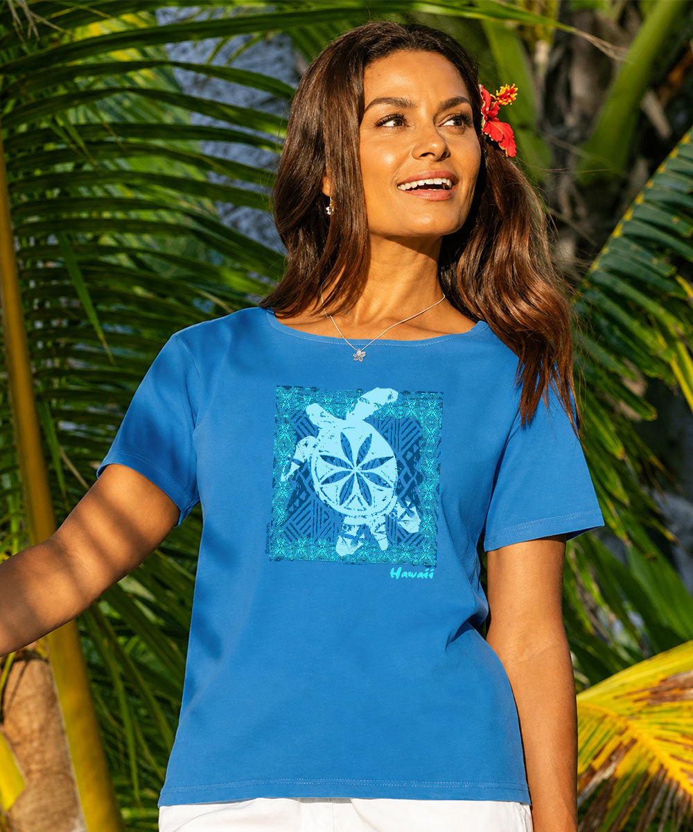 A woman wearing a bright blue short-sleeve t-shirt with a blue and white Hawaiian turtle graphic on the front, posing outdoors in front of tropical palm leaves. View 2