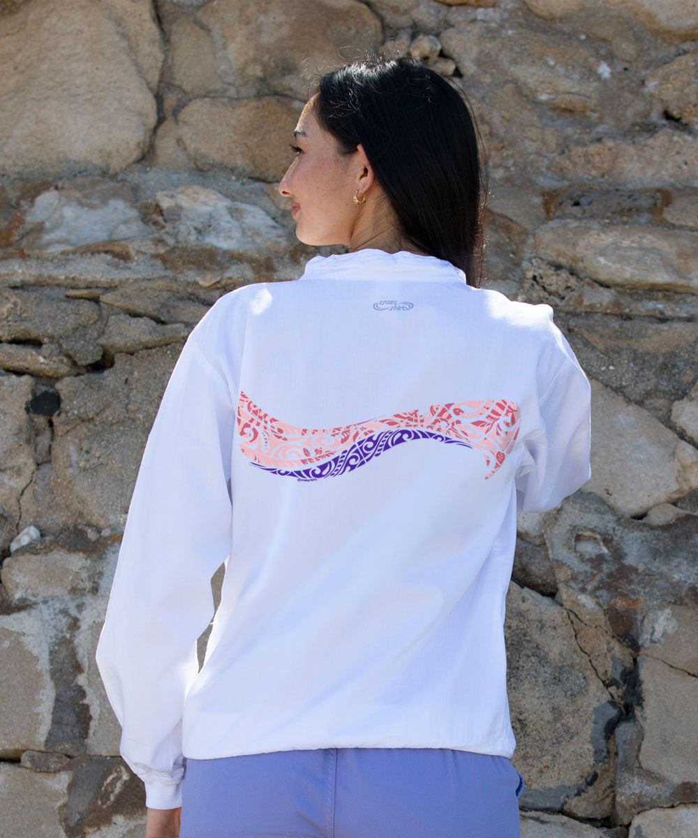A woman viewed from behind wearing a white long-sleeve athletic jacket with a wavy graphic across the back in pink and purple, paired with lavender pants, standing against a rustic stone wall. View 3
