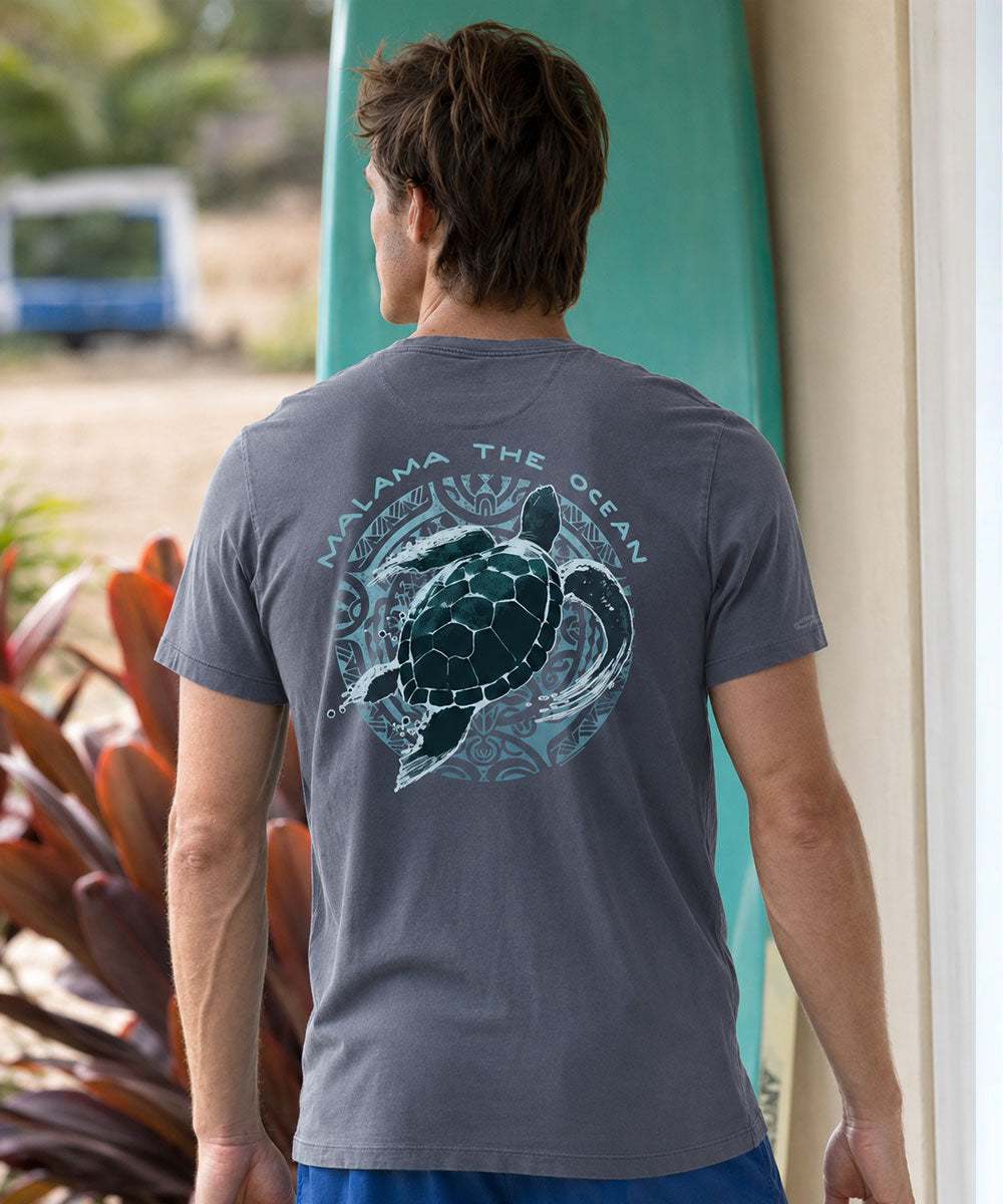 A man viewed from behind wearing a dark gray t-shirt with a large graphic of a sea turtle and the text 'MALAMA THE OCEAN' on the back. He is standing next to a teal surfboard in a beachside setting. View 2