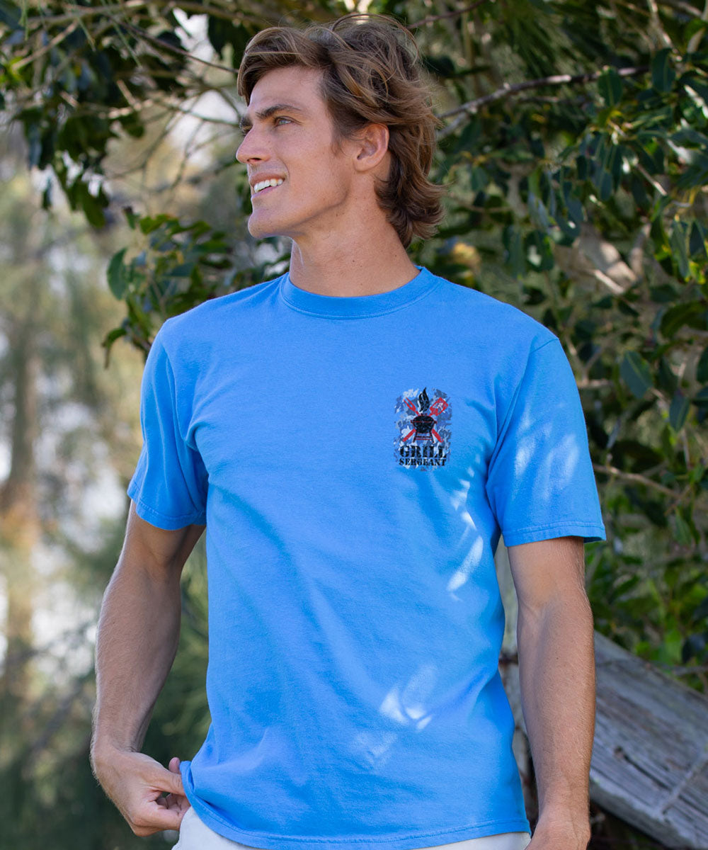 A man wearing a bright blue crewneck t-shirt with a small graphic on the left chest, standing outdoors in front of a leafy green background. View 3