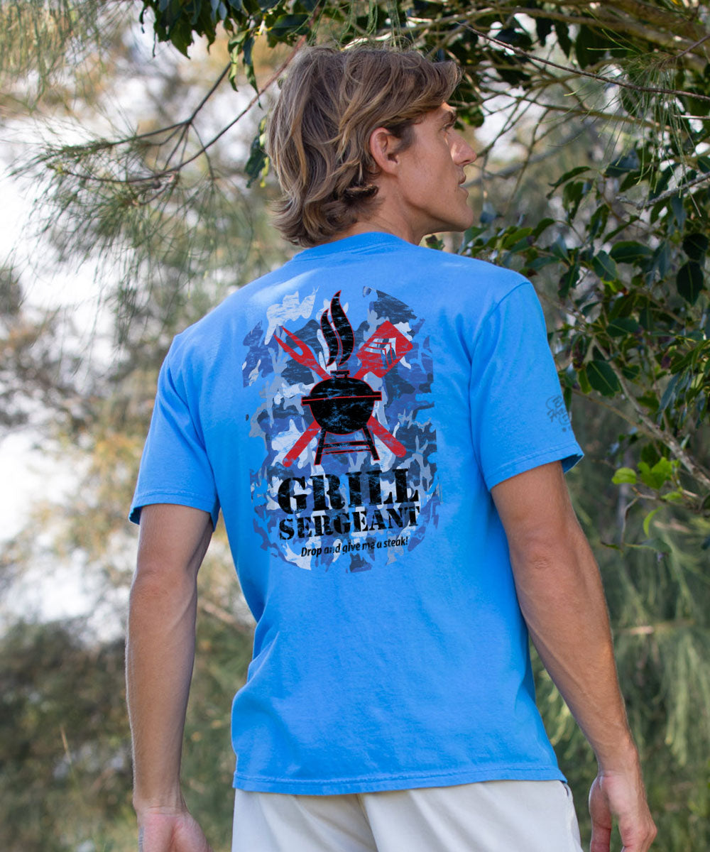 A man wearing a bright blue t-shirt with a large graphic on the back reading 'GRILL SERGEANT' photographed outdoors in front of trees. View 2