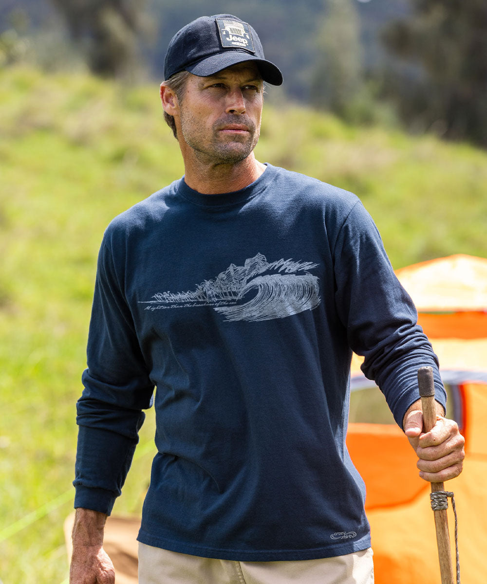 A man wearing a navy blue long-sleeve t-shirt with a white wave graphic on the chest, a navy baseball cap, and khaki pants, holding a wooden pole outdoors in a grassy field. View 2