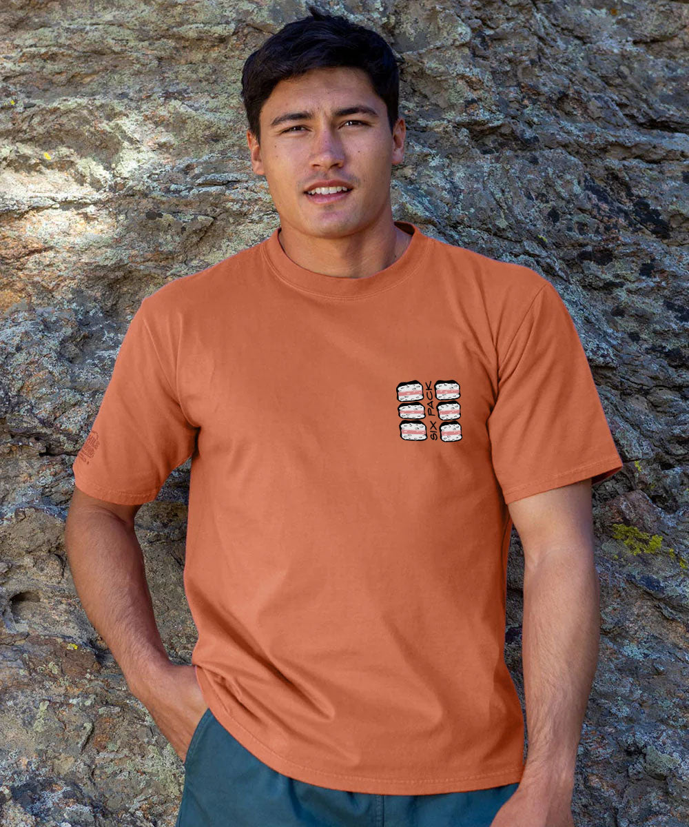 A man wearing a terracotta orange crewneck t-shirt with a small graphic print on the left chest, standing against a textured rock wall. View 3