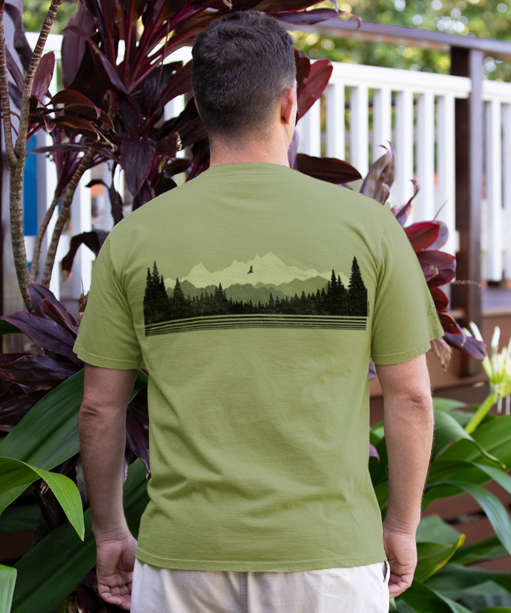 A man viewed from behind wearing an olive green t-shirt with a black graphic of a mountain range and pine forest across the upper back, set against a background of tropical plants and a white railing. View 3
