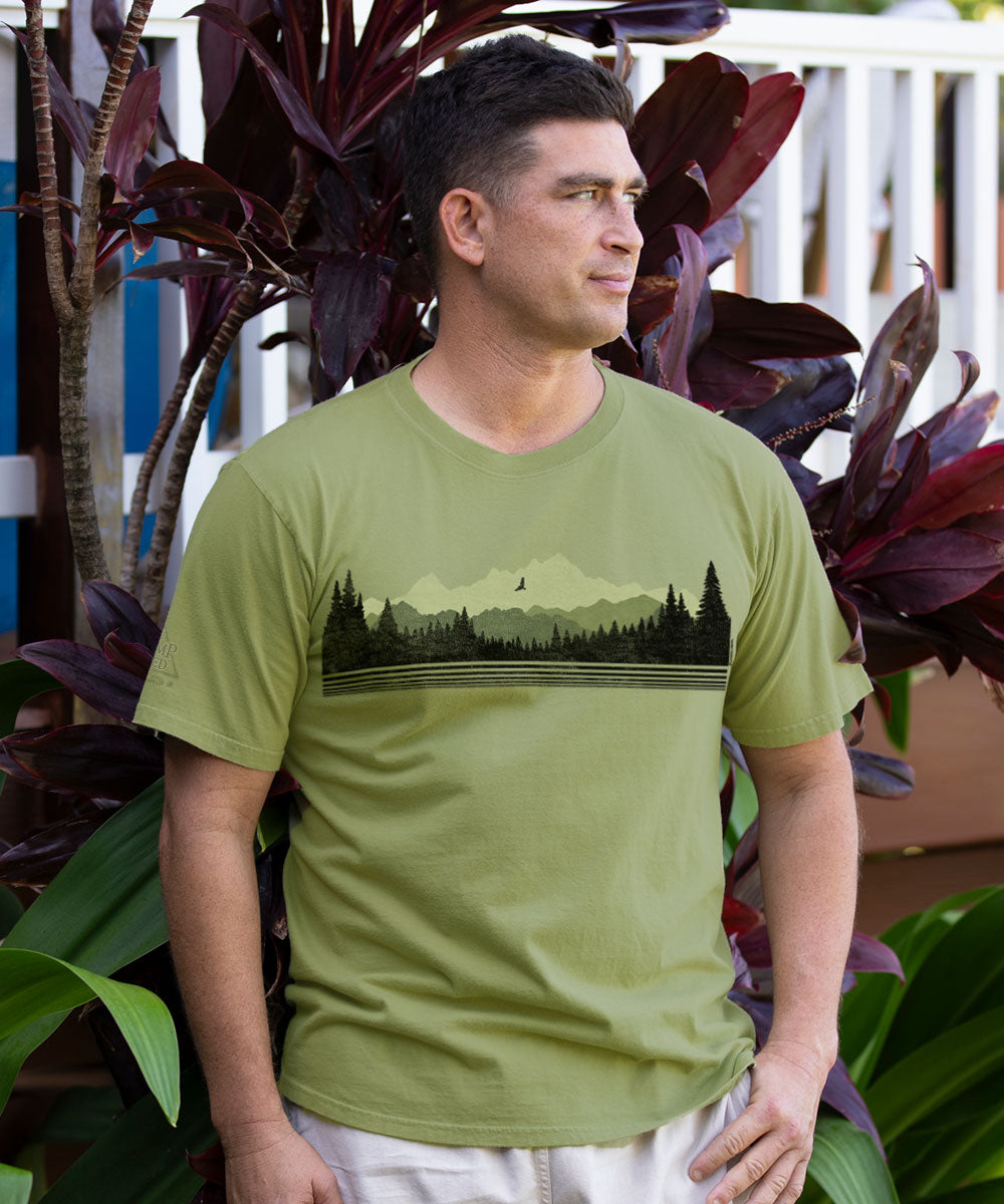 A man wearing an olive green t-shirt with a black forest and mountain landscape graphic across the chest, standing in front of tropical plants. View 2