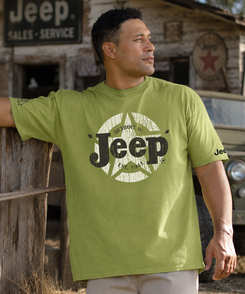 Man wearing a green Jeep t-shirt leaning against a wooden fence with trees in the background.
