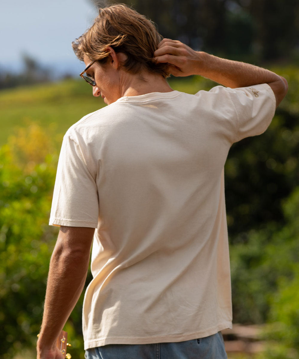 Man in a beige t-shirt standing outdoors with greenery in the background View 3