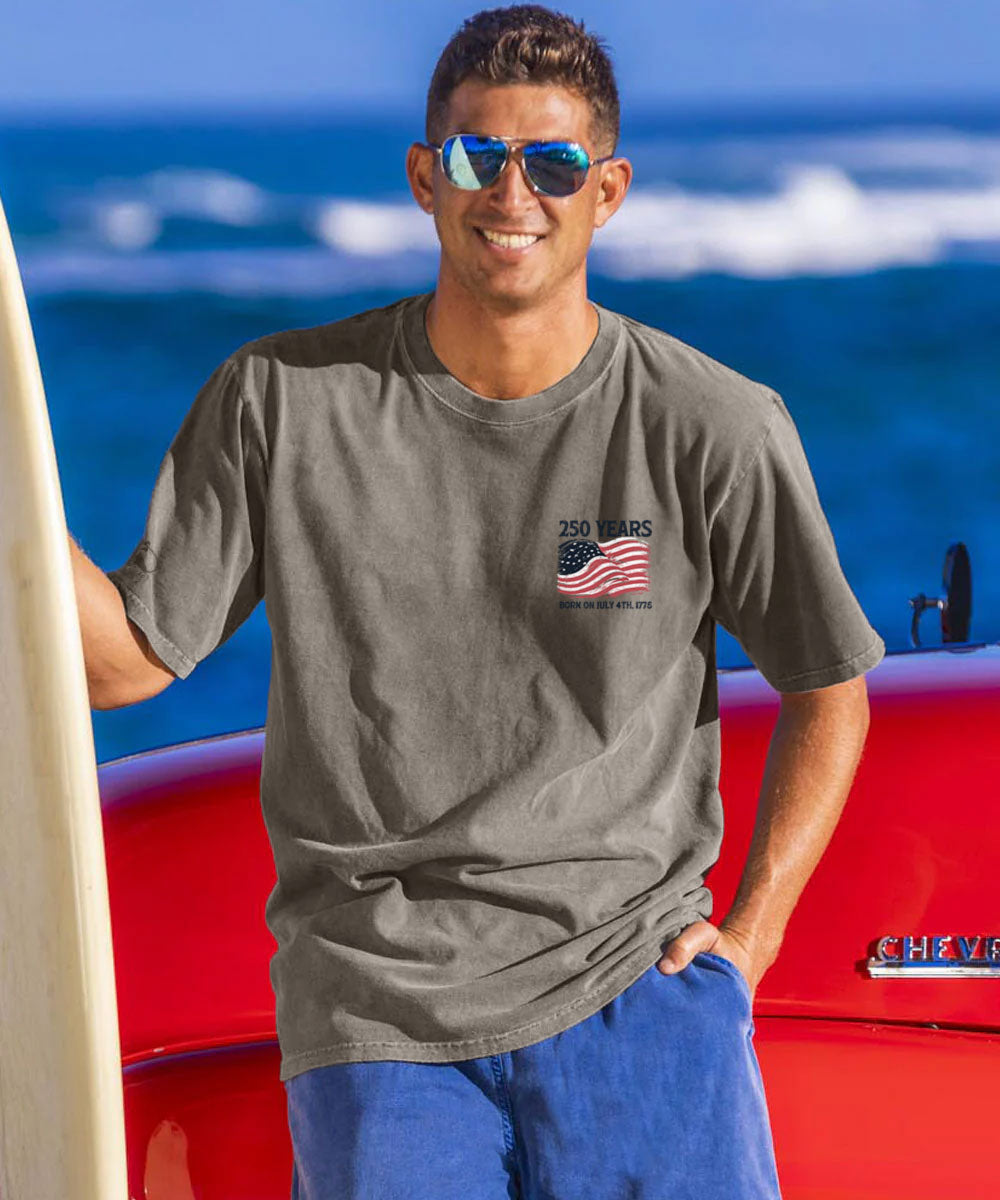 Man wearing a gray t-shirt with a logo, sunglasses, and blue shorts standing next to a red car with ocean in the background View 3