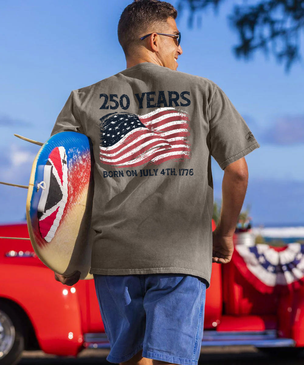 Man wearing a t-shirt with '250 Years Born on July 4th, 1776' design, holding a shield with American and British flags, standing in front of a red truck.
